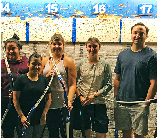 Five people standing indoors in front of an archery target wall, smiling at the camera and holding bows—like a winning team all about Abby Connect. Numbered targets 14 to 17 are visible above their heads.