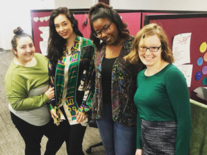 Four women stand together in an office space at Abby Connect, smiling at the camera. The background shows maroon partition walls decorated with papers and colorful circles. They are wearing colorful, casual outfits.